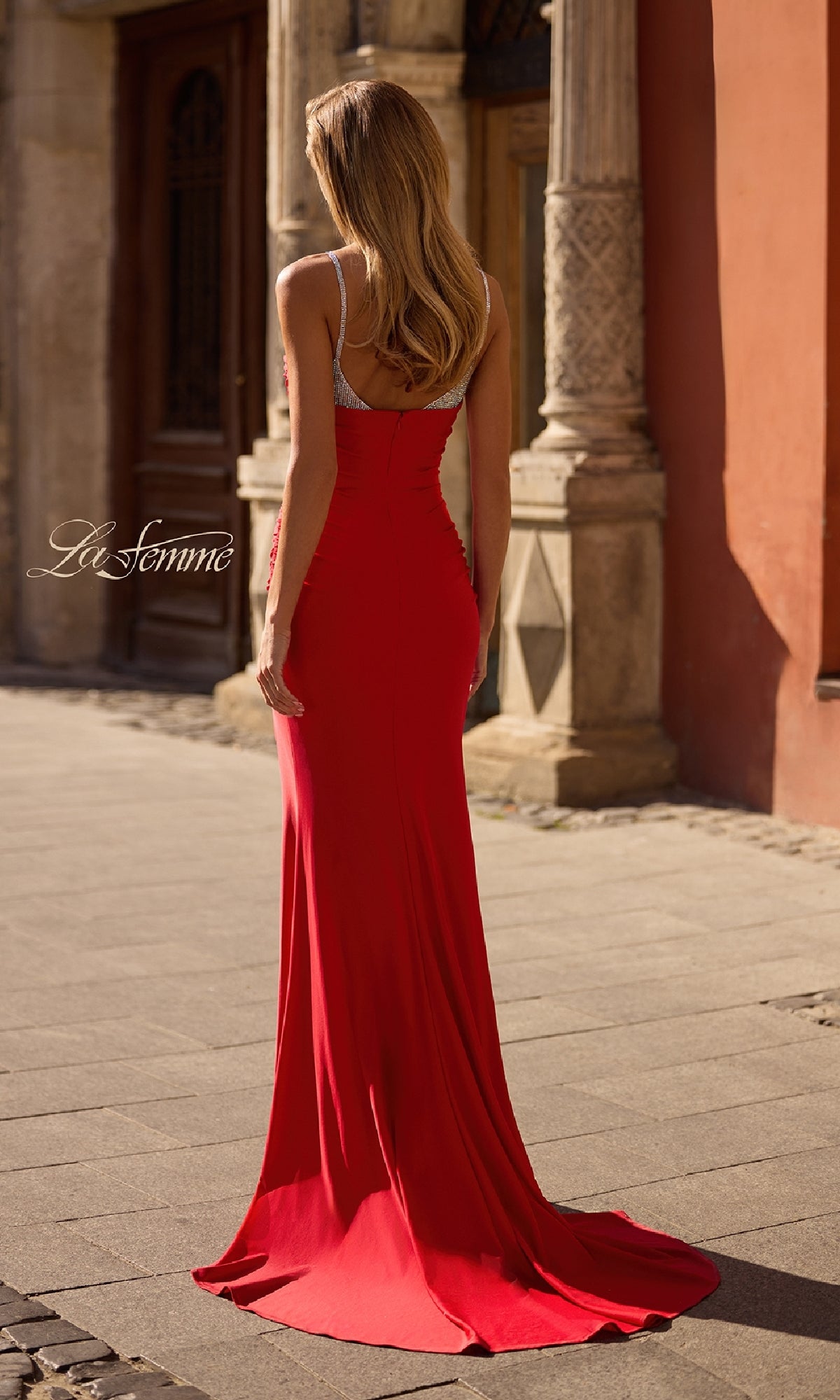 A woman with long hair stands outdoors in the La Femme 32939 Beaded-Neckline Long Prom Dress, featuring thin straps and a beaded v-neckline, facing away from the camera.
